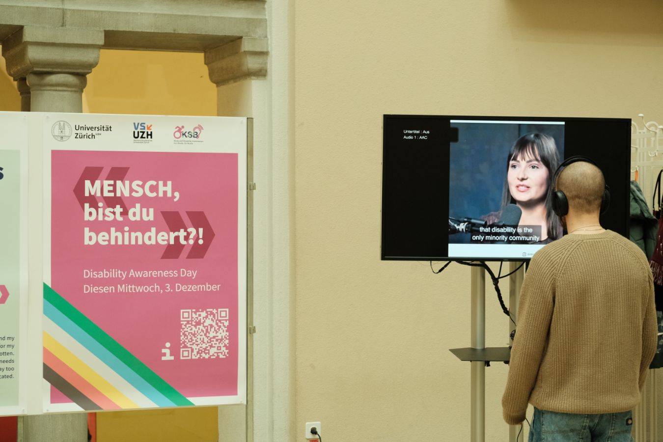 Ausstellung Videos: Eine Person steht vor einem Bildschirm mit einem Video, das Untertitel zeigt; daneben ein Plakat zum „Disability Awareness Day“.